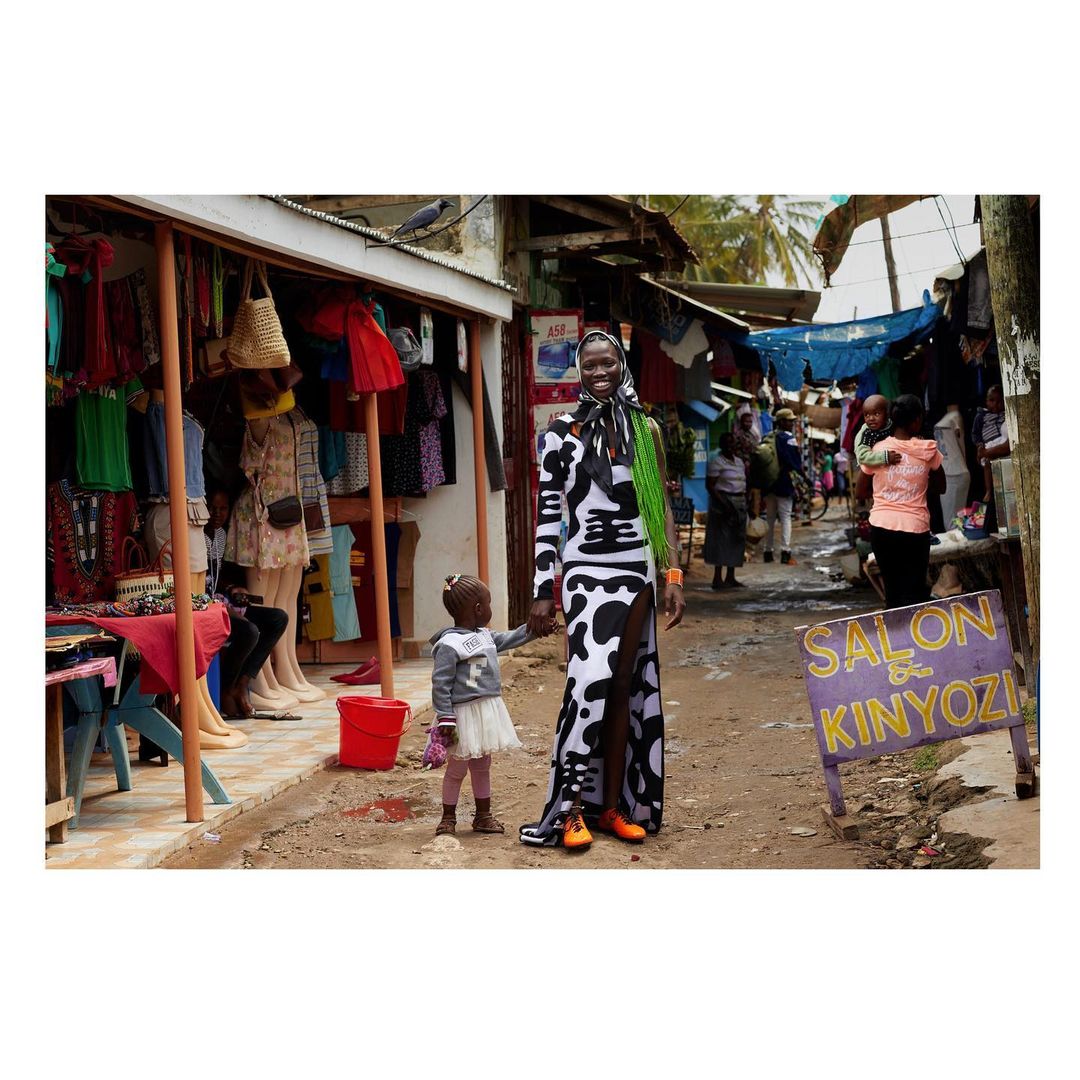A woman and child stand in a marketplace in bright sunlight. The woman wears a split black and white dress and bright orange shoes.