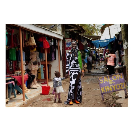 A woman and child stand in a marketplace in bright sunlight. The woman wears a split black and white dress and bright orange shoes.