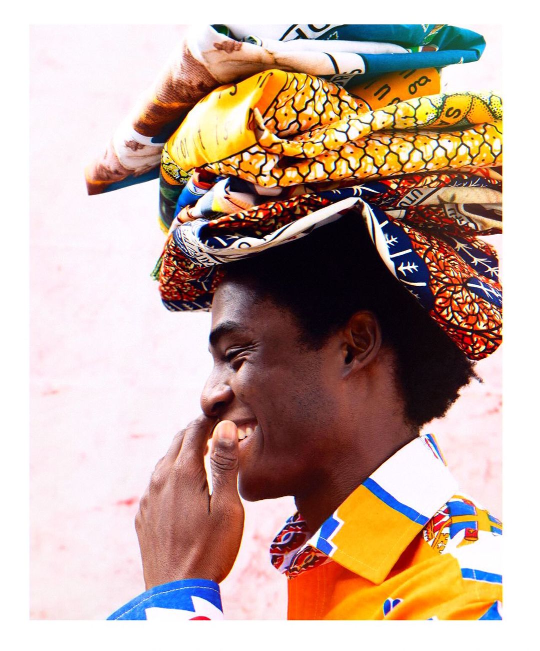 Profile shot of a Black adult male with patterned textiles balanced on his head against a pale pink background, smiling widely.