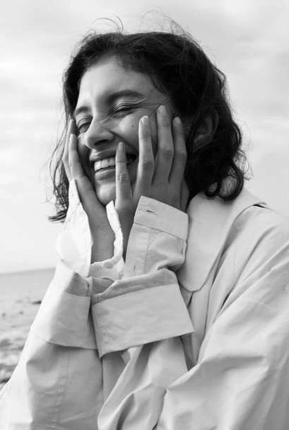Black and white portrait of a smiling woman with hands on her face, wearing a buttoned shirt or jacket, with a blurred outdoor background.