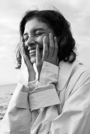Black and white portrait of a smiling woman with hands on her face, wearing a buttoned shirt or jacket, with a blurred outdoor background.