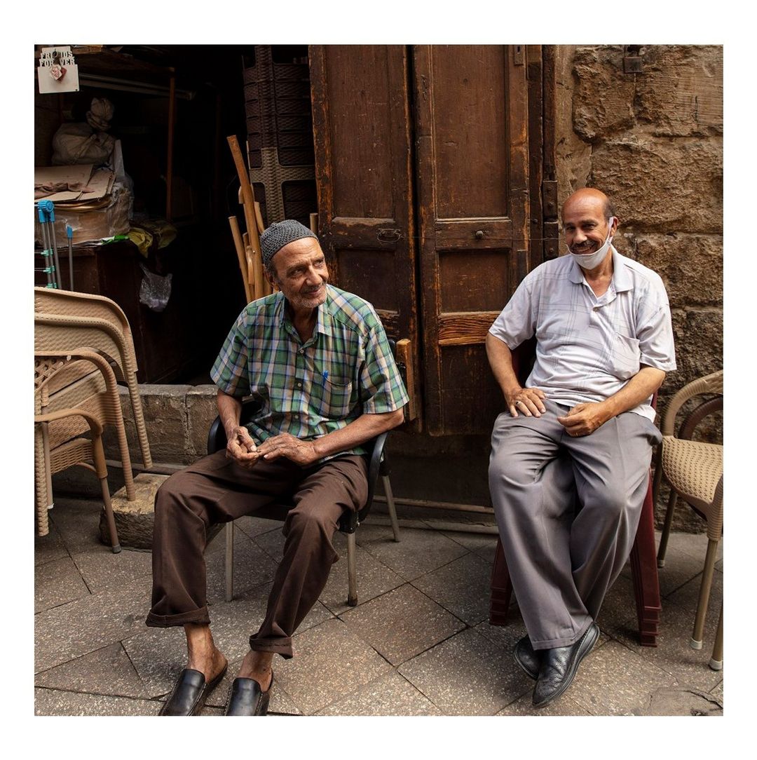 Two elderly men seated on chairs outside a building, one wearing a mask, creating a candid, casual scene with textured details.