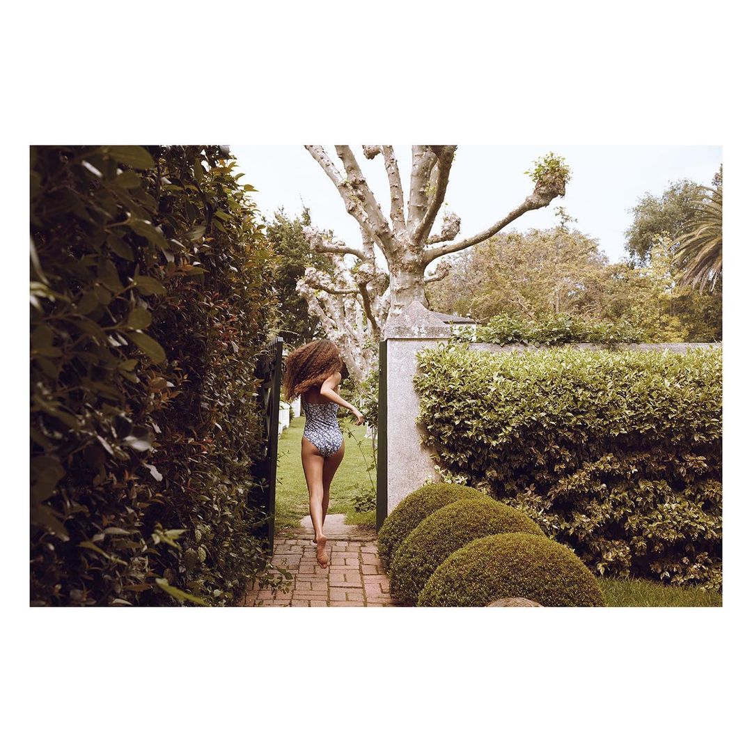 A woman with curly hair in a swimsuit runs on a brick path through a garden with green hedges and trees on a sunny day outdoors.