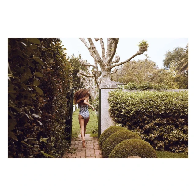 A woman with curly hair in a swimsuit runs on a brick path through a garden with green hedges and trees on a sunny day outdoors.