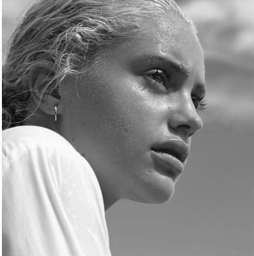 Monochrome portrait of a blonde adult female with short hair, wearing a white shirt, and earring, set against a cloudy blue background.