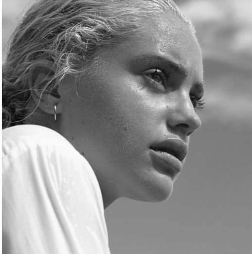 Monochrome portrait of a blonde adult female with short hair, wearing a white shirt, and earring, set against a cloudy blue background.