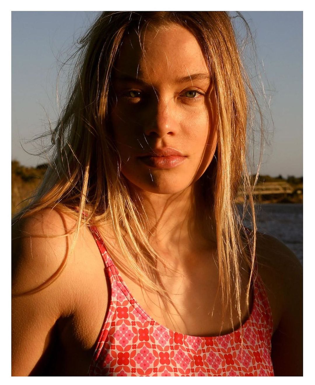 Portrait of a young adult woman with blonde hair and fair skin wearing a floral top, standing outdoors with a natural background.