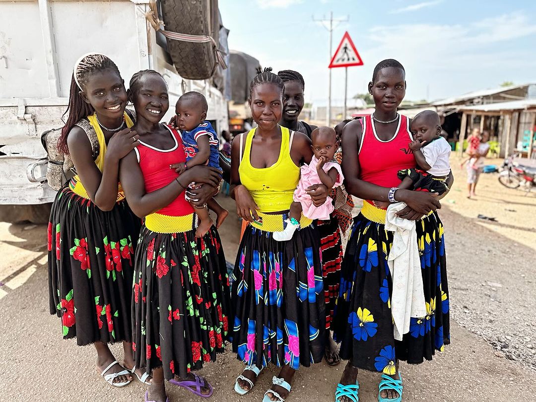 Group of women and children in colorful traditional clothing pose outdoors near a dirt road and village, with a vehicle in the background.