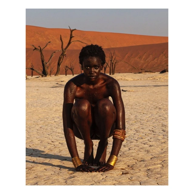African woman squats in a desert landscape wearing gold bracelets in the Namib desert against a sand dune background with dead trees.