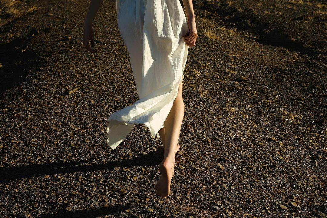 Barefoot woman in white dress walks across arid, rocky landscape in golden light. Low angle captures movement and natural textures.