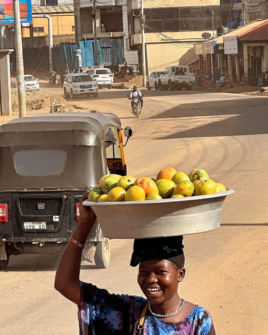 Smiling African woman carrying a basin of fruit on her head on a dusty urban street with cars and a motorcycle in the background.