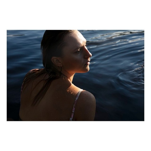 Woman partially submerged in water, facing away from the camera. The light catches her skin and wet hair, creating a serene scene.