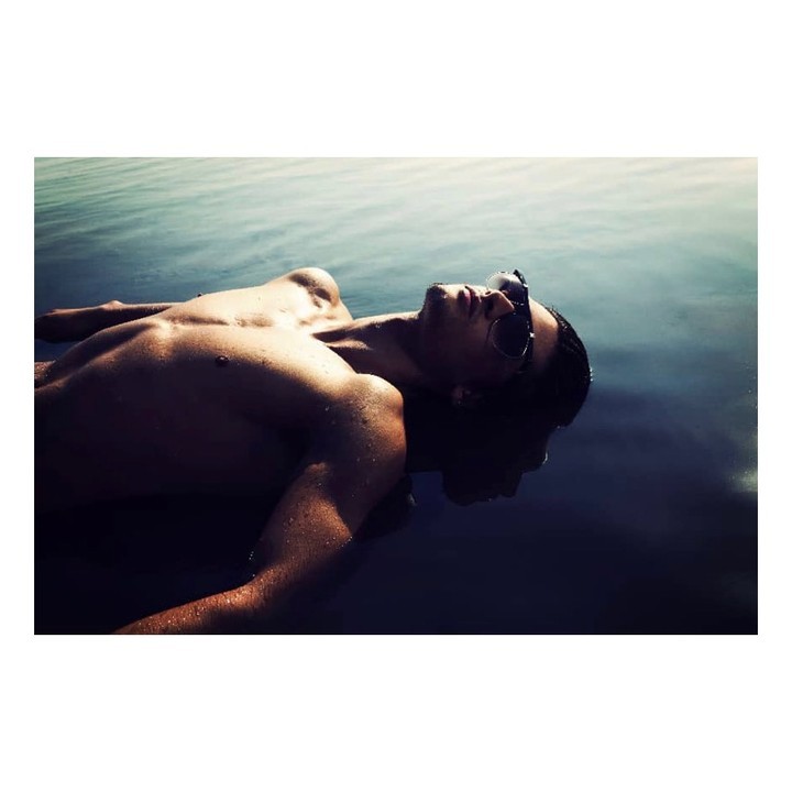 A shirtless adult male lies on his back in water wearing sunglasses. The water is dark with some visible ripples and reflections.