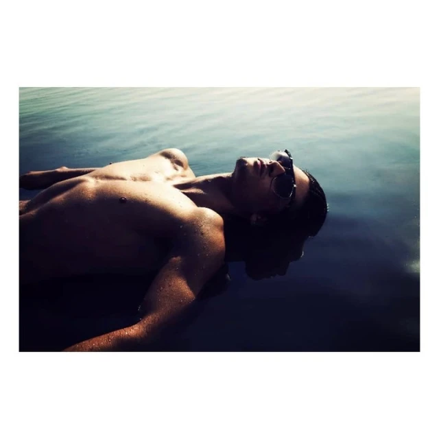 A shirtless adult male lies on his back in water wearing sunglasses. The water is dark with some visible ripples and reflections.