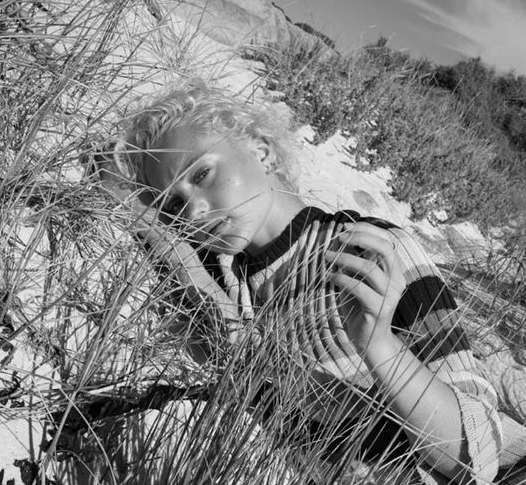 Black and white portrait of a young adult woman laying in tall grass outdoors on a sunny day with a close-up, eye-level perspective.
