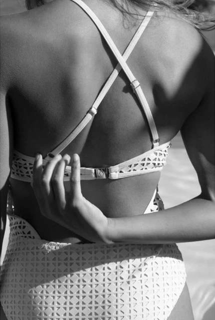 Back view of an adult woman in a white lattice-patterned swimsuit with crossed straps, adjusting the back closure in an outdoor setting.