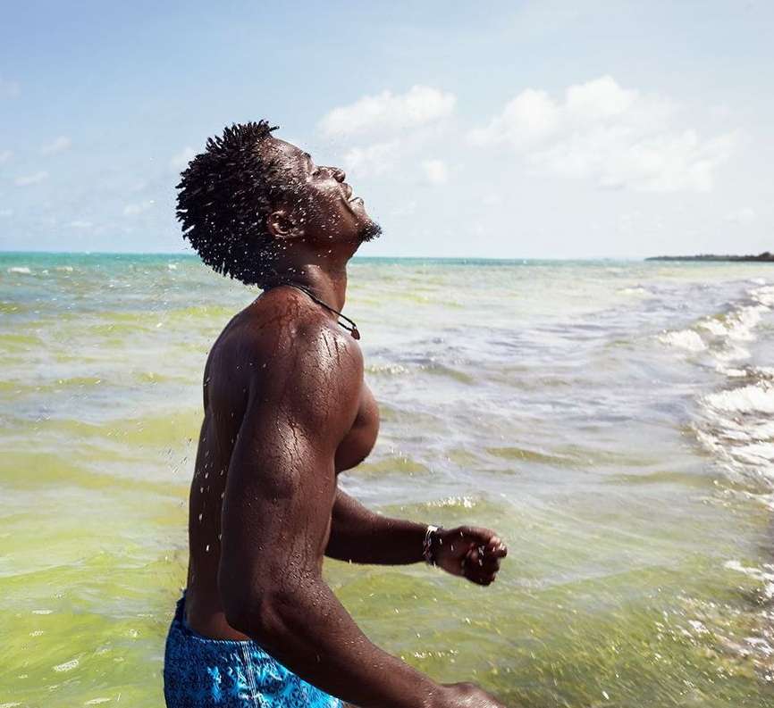 Athletic man in blue shorts standing in shallow ocean water with head tilted back and eyes closed; splashing water, bright sunlight, clear sky.