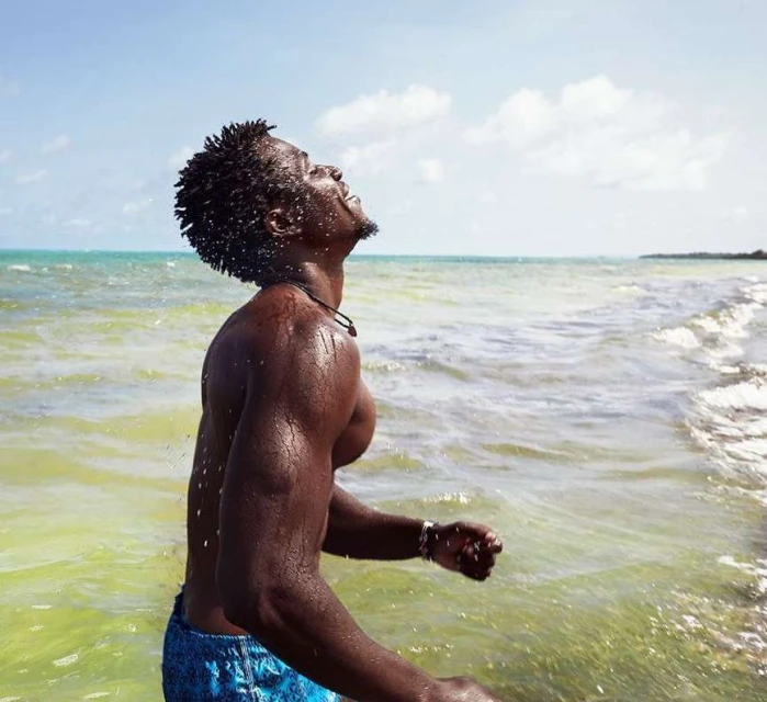 Athletic man in blue shorts standing in shallow ocean water with head tilted back and eyes closed; splashing water, bright sunlight, clear sky.