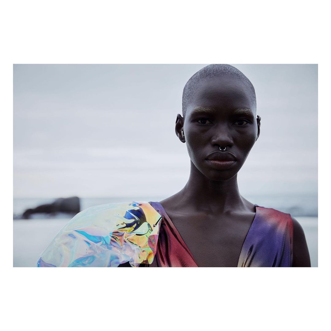 Close-up of a Black woman with a shaved head wearing a colorful, iridescent dress, posing in front of a muted beach landscape with grey skies.