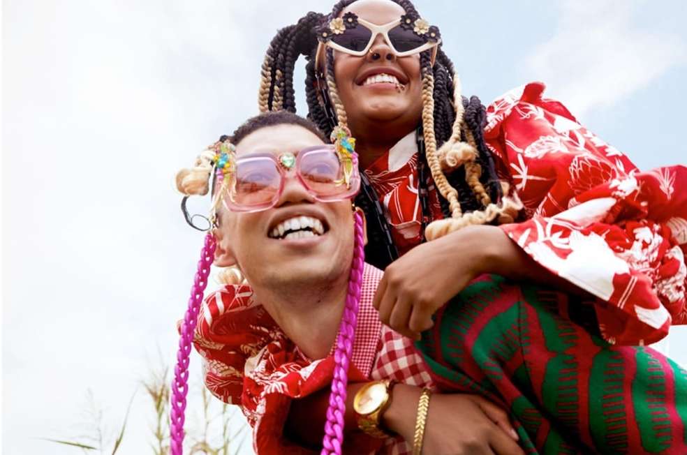A happy couple posing outdoors in fashionable clothes and funky sunglasses, with a cloudy sky in the background, and both are smiling.