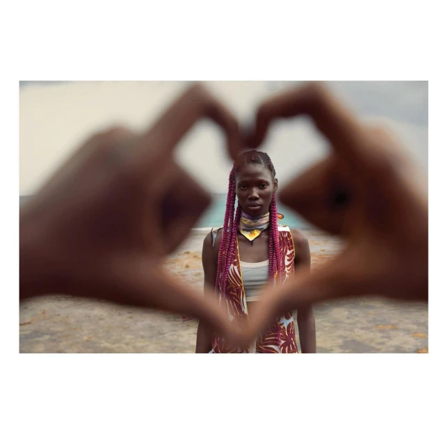 Portrait of a dark-skinned woman with pink braids making a heart shape with her hands in front of a beach landscape, summer fashion.