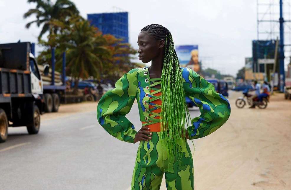 Woman in green patterned two-piece outfit and bright green braids standing on a road with trucks, cars, and buildings in the background.