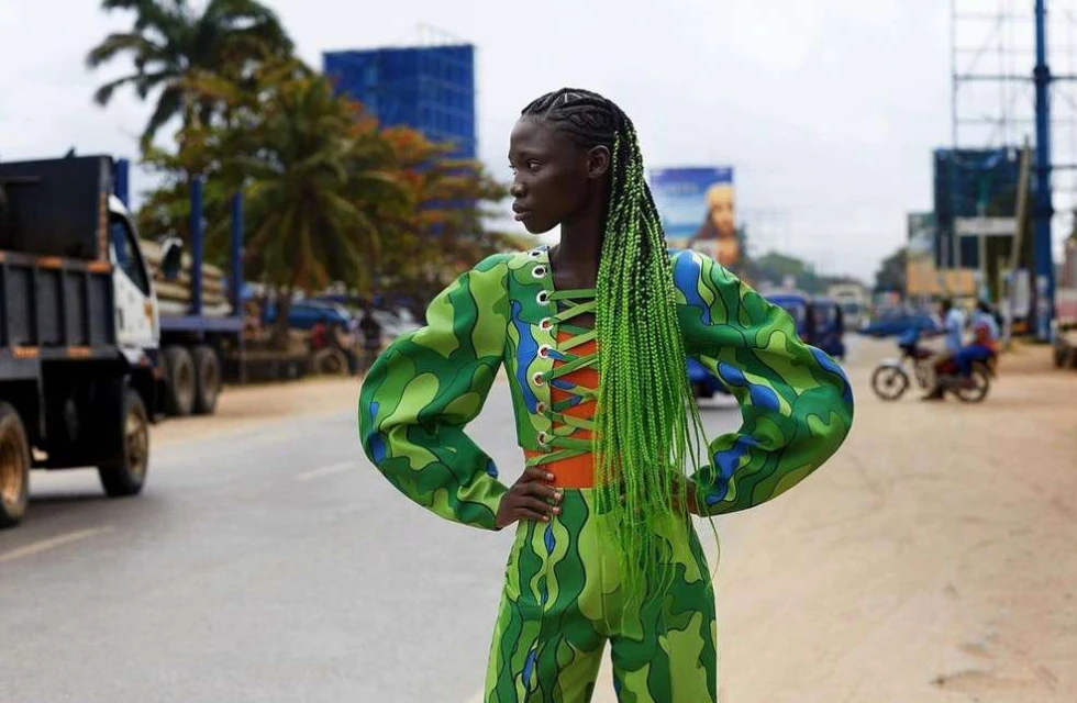 Woman in green patterned two-piece outfit and bright green braids standing on a road with trucks, cars, and buildings in the background.