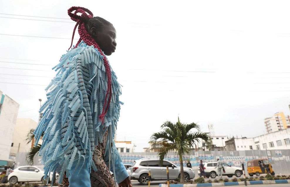 Adult female in an urban setting wearing a fringe jacket and braided hair with city buildings and palm trees visible in the background.