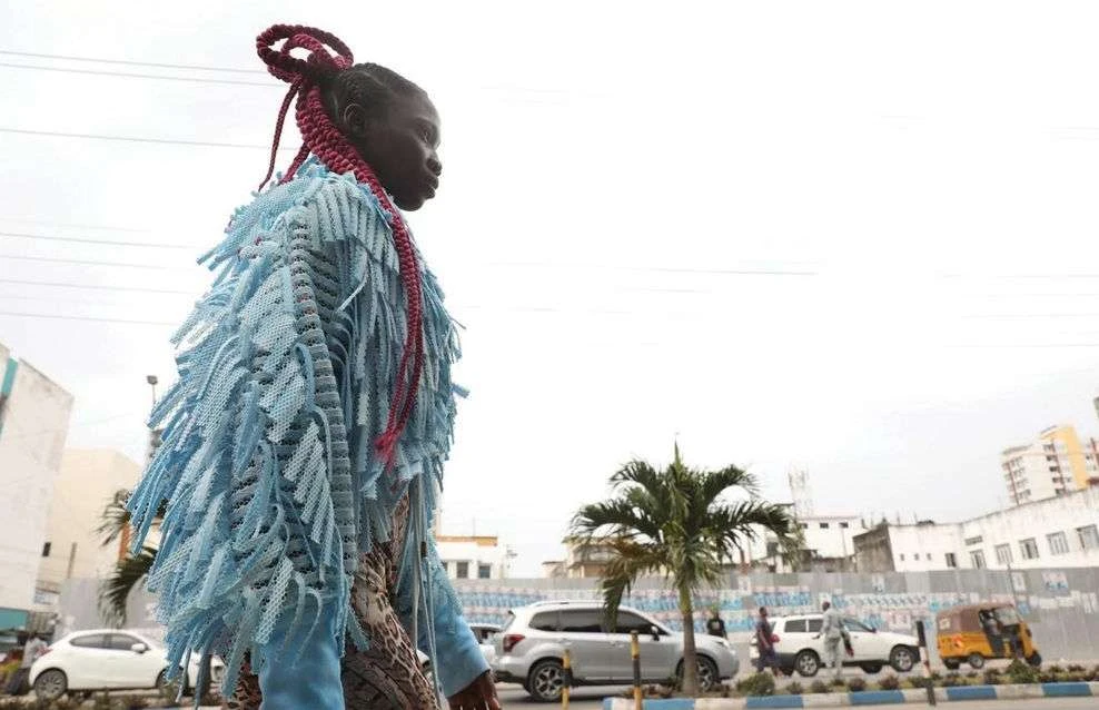 Adult female in an urban setting wearing a fringe jacket and braided hair with city buildings and palm trees visible in the background.