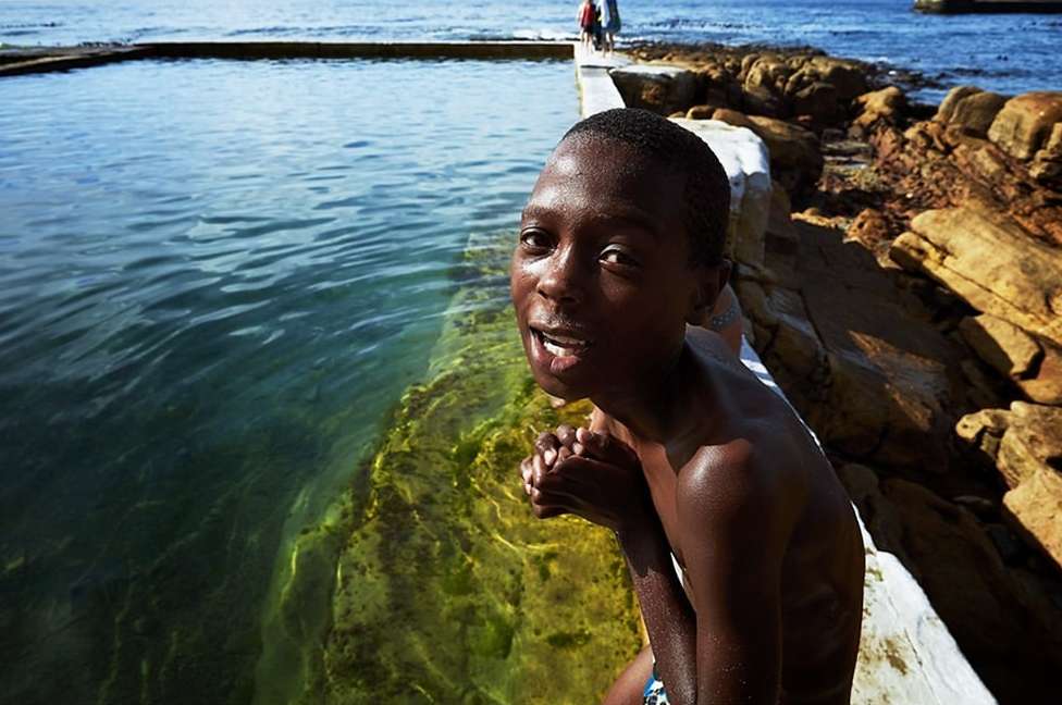 A smiling Black child sits on the edge of a concrete pool with rocky cliffs and blue ocean water visible in the background on a sunny day.