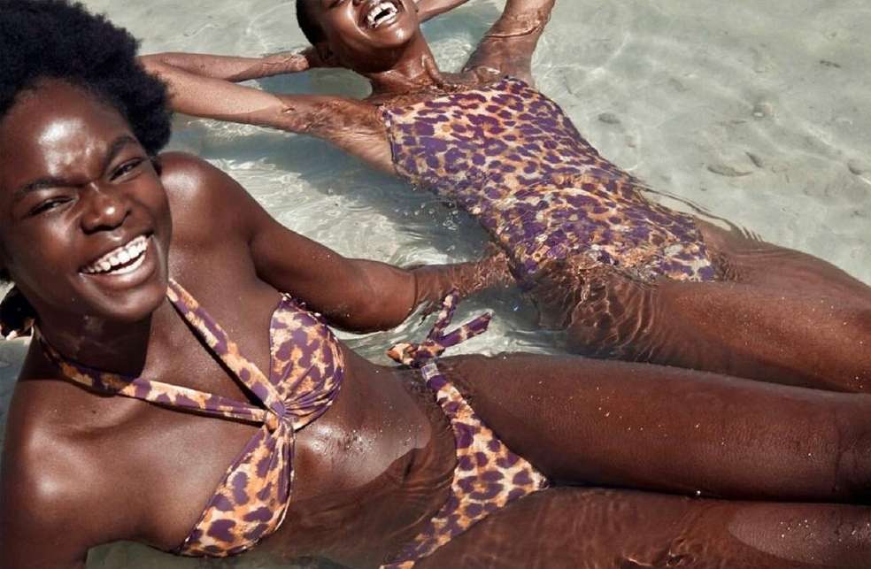 Two smiling women wearing animal print swimwear relax in shallow, clear water near a sandy beach. Bright, sunny, summertime mood.