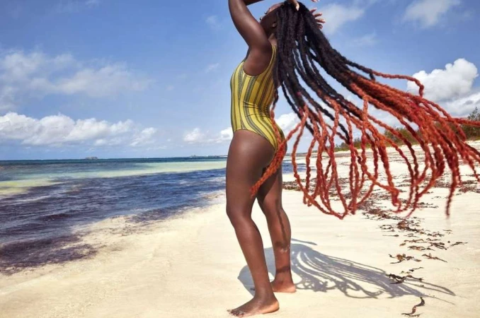 Black woman with very long dreadlocks wearing a yellow striped one-piece swimsuit on a sunny sandy beach