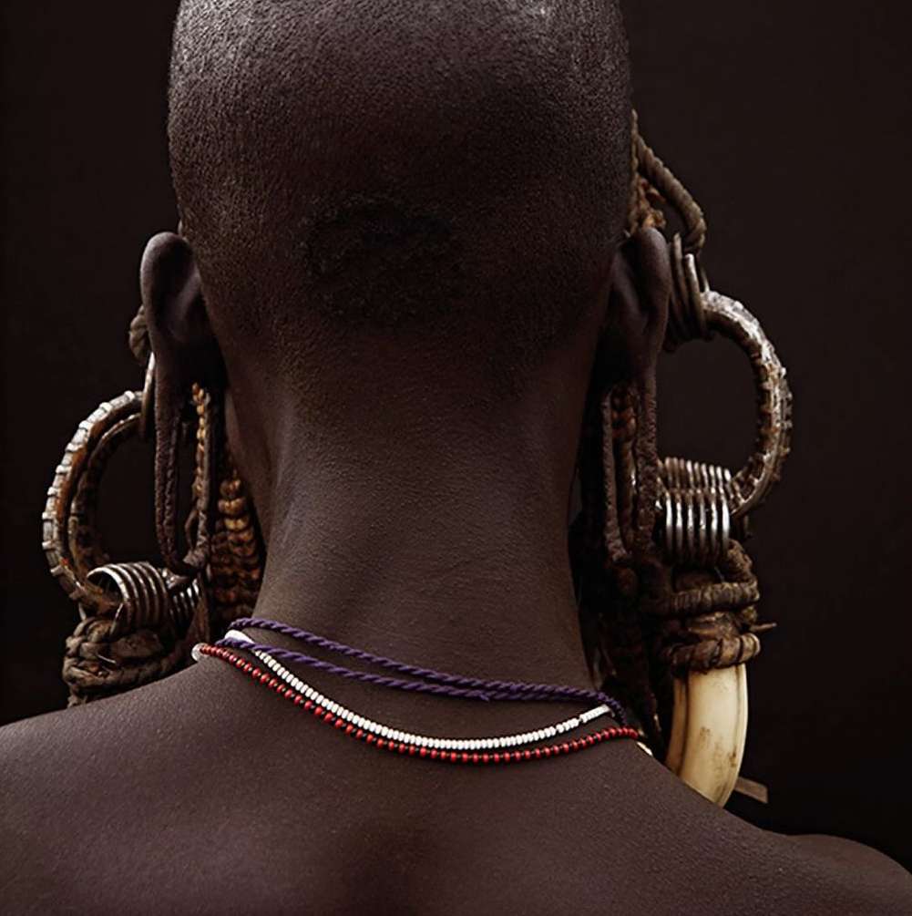 Close-up studio shot of an adult with dark skin, large earrings, and necklaces against a dark background, showcasing cultural adornment.