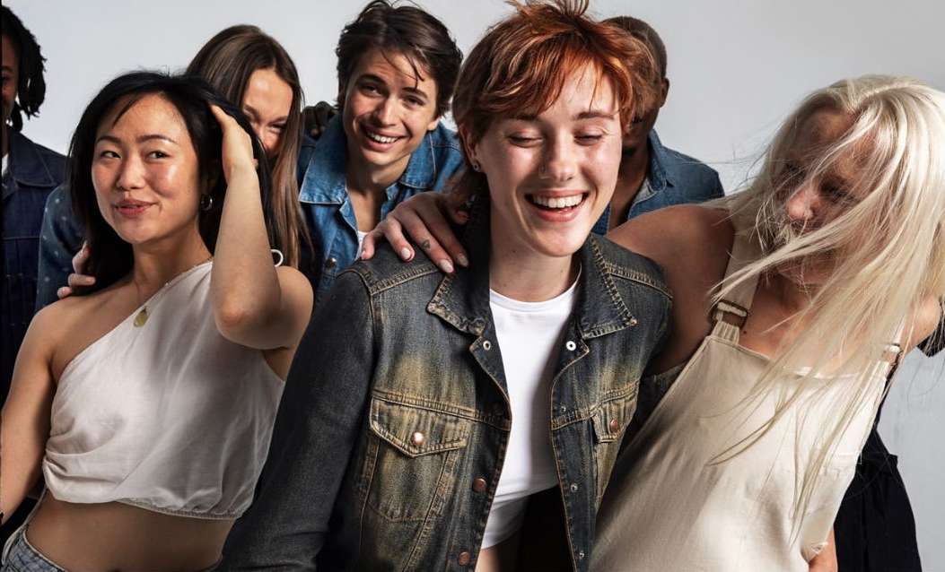 A group of casually dressed, diverse young adults are closely grouped together in an eye-level studio shot against a white backdrop.