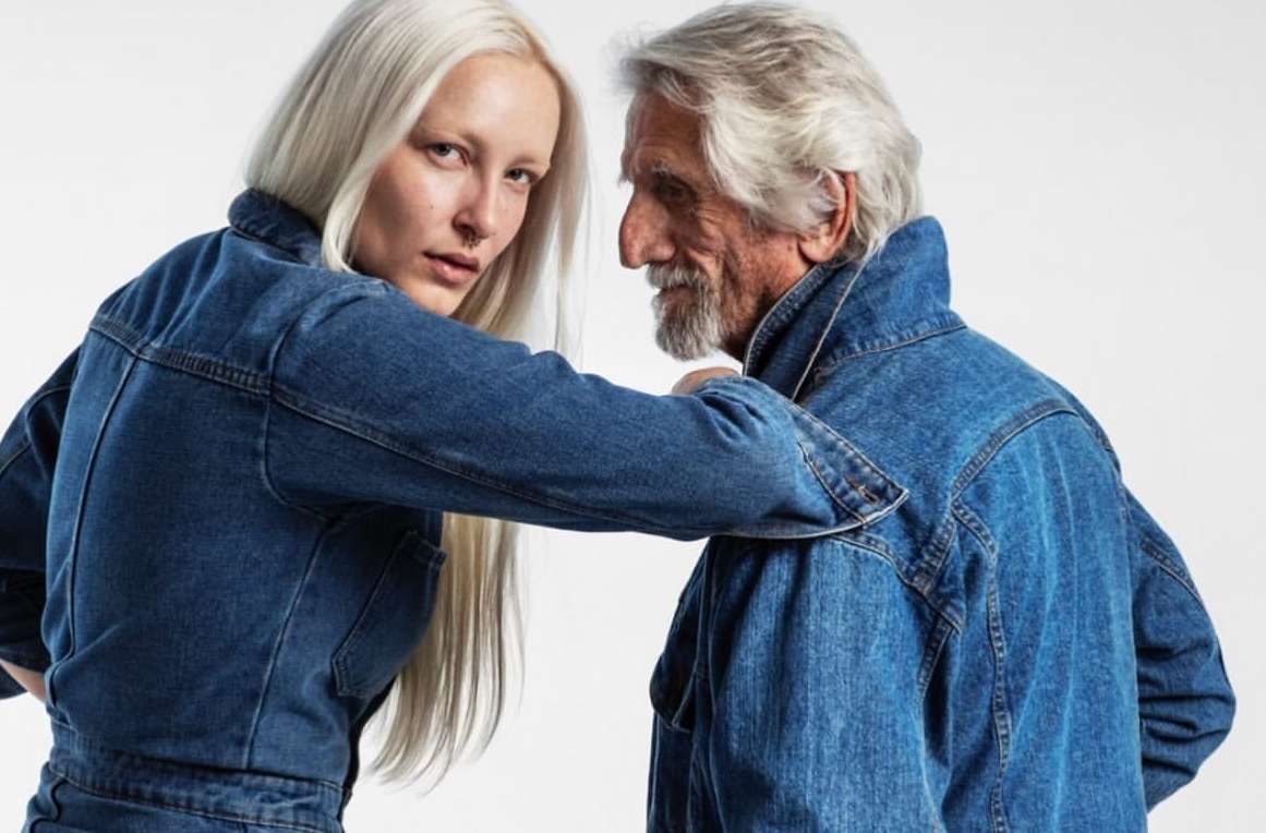 Denim fashion portrait: young adult woman with platinum hair and an elderly man in matching jean jackets against a white backdrop.
