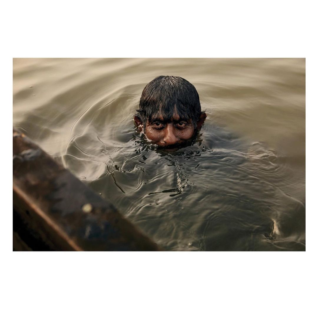 Adult male submerged in water up to his neck, looking directly at the camera in an outdoor setting with overcast lighting.