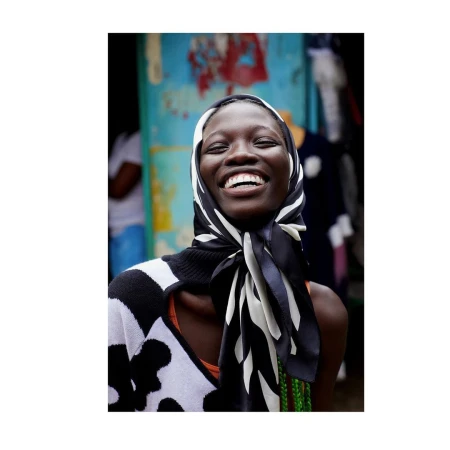 Smiling African woman in a patterned scarf and sweater stands outdoors with a joyful expression, showcasing a vibrant and fashionable portrait.