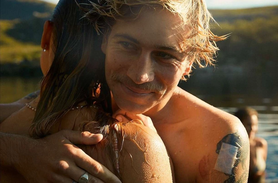 Close-up of a smiling man with wet hair and a mustache hugging a woman in a lake setting, with a bright sunny day and visible tattoos.