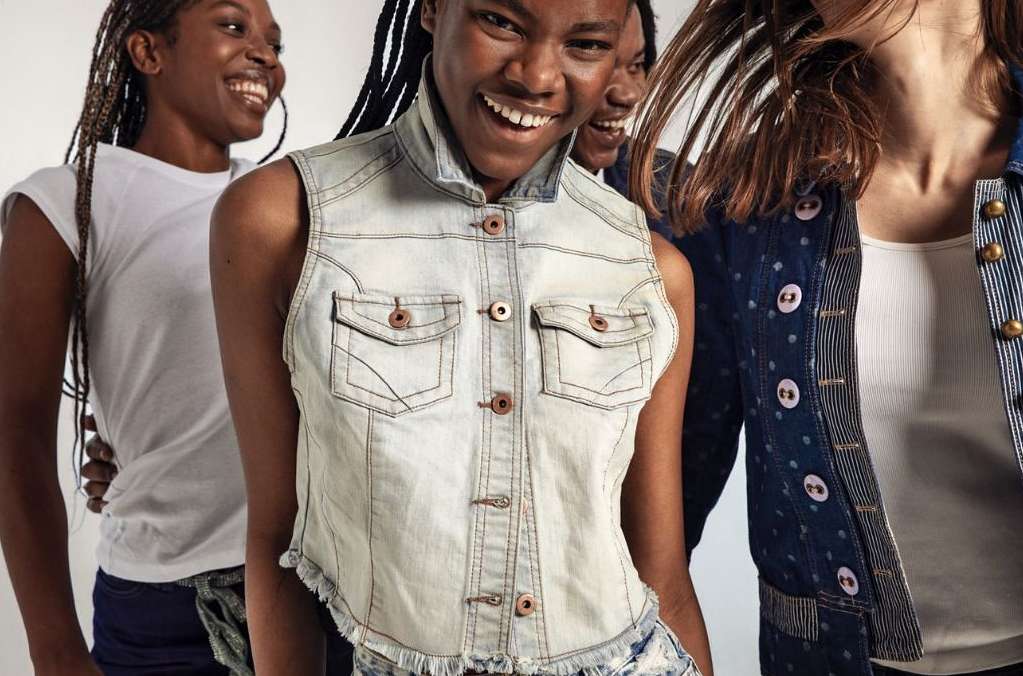 Three smiling Black women in light denim and white T-shirts pose indoors in a bright studio. Fashionable and modern clothing styles.
