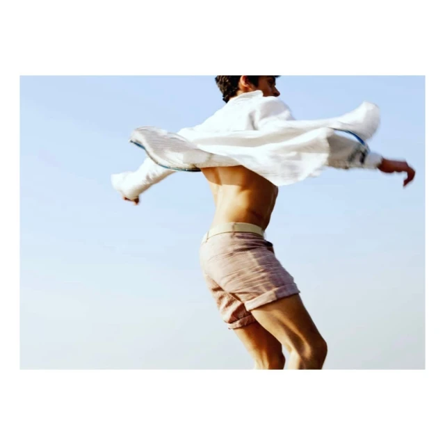 Athletic adult male in tan shorts and white shirt poses with arms out against a clear blue sky in an outdoor fashion shot.