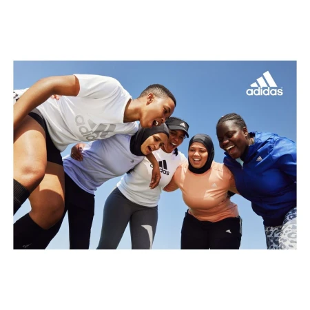 A diverse group of smiling women in activewear, some wearing hijabs, gather outdoors under a bright blue sky with the Adidas logo.