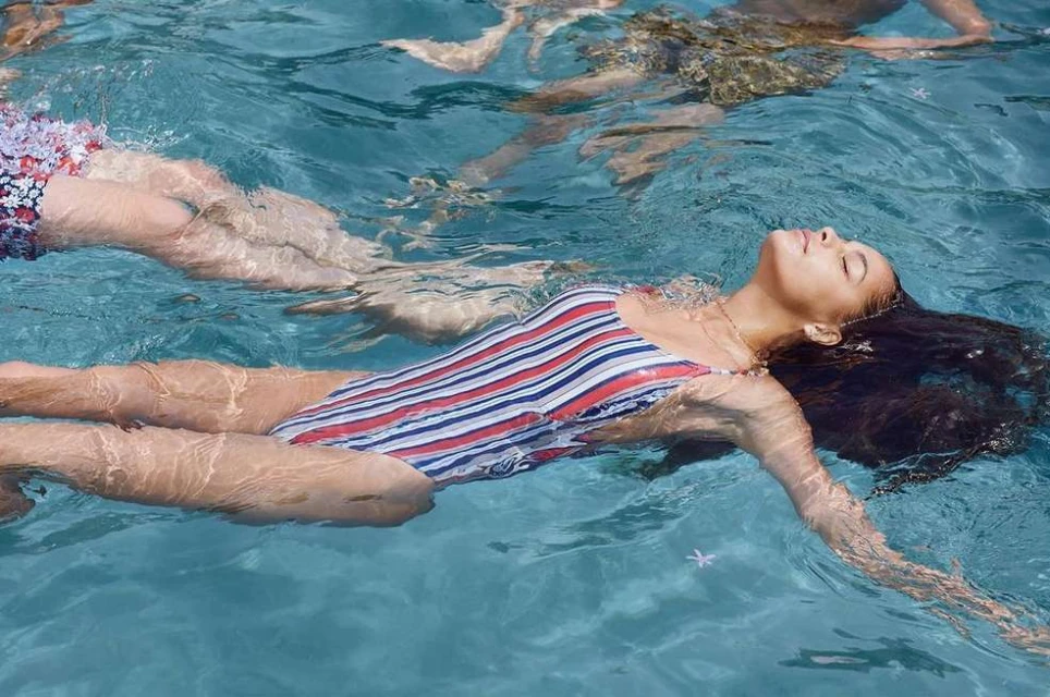 Woman floats in swimming pool with red, white, and blue striped swimsuit. Relaxing pose with closed eyes in blue water.