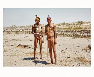 Two Black men holding wooden spears standing on a sandy ground with zebras in the background by photographer Alexa Singer.