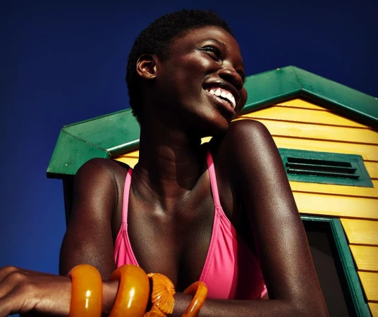 Smiling black woman wearing a pink bikini top and chunky amber bangles against a yellow beach hut and deep blue sky.