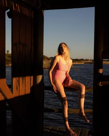 Woman in patterned pink swimsuit seated on a wooden railing outdoors by water during sunset by Alexa Singer