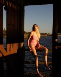 Woman in patterned pink swimsuit seated on a wooden railing outdoors by water during sunset by Alexa Singer