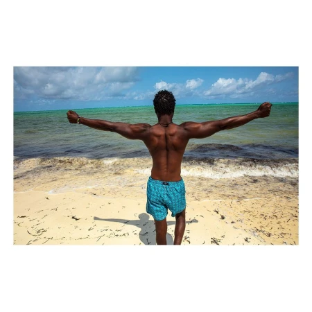 Shirtless man wearing blue patterned swim shorts standing on a sandy beach with arms outstretched toward the ocean, captured from behind.