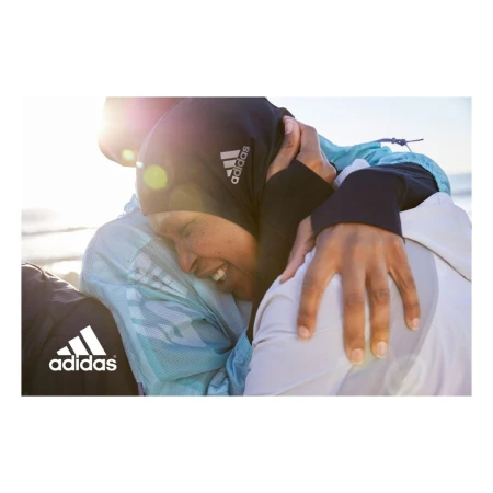 Two women hugging outdoors wearing sports apparel with visible Adidas branding against a bright sunny sky background.