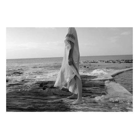 Person standing on rocky coastal shore wearing a long patterned tunic by the sea in black and white photography.