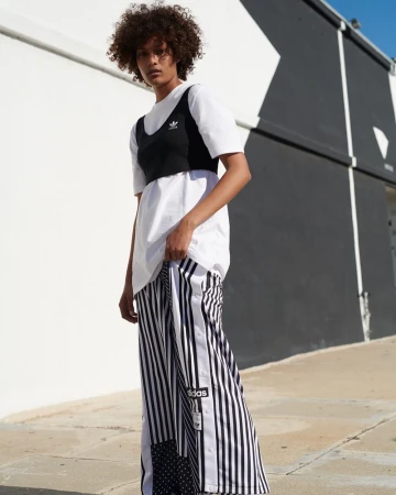 A woman with curly hair poses against a black-and-white wall wearing a white shirt, black crop top, and striped Adidas track pants outdoors.